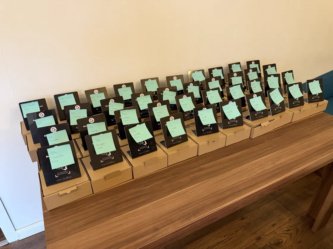 Gepackte Versandkartons mit schwarzen NFC-Aufstellern, auf jedem ein Zettel „Bitte Schutzfolie abziehen“