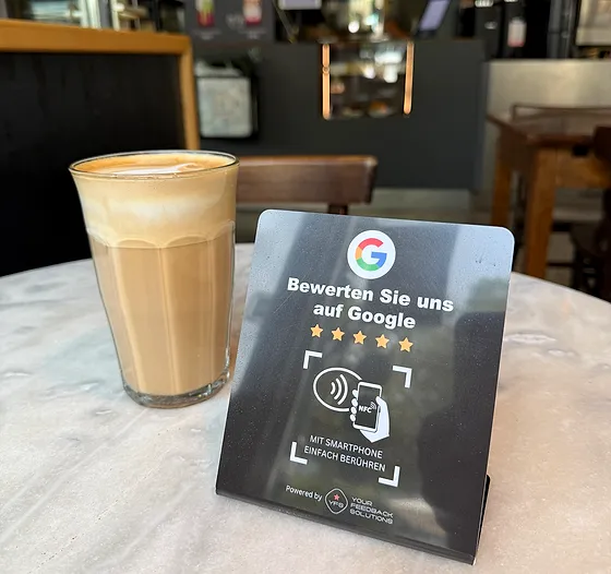Der NFC-Aufsteller steht auf einem Marmortisch in einem Café, daneben ein Milchkaffee im Glas; im Hintergrund Tresen, Siebträgermaschine und Menütafeln.